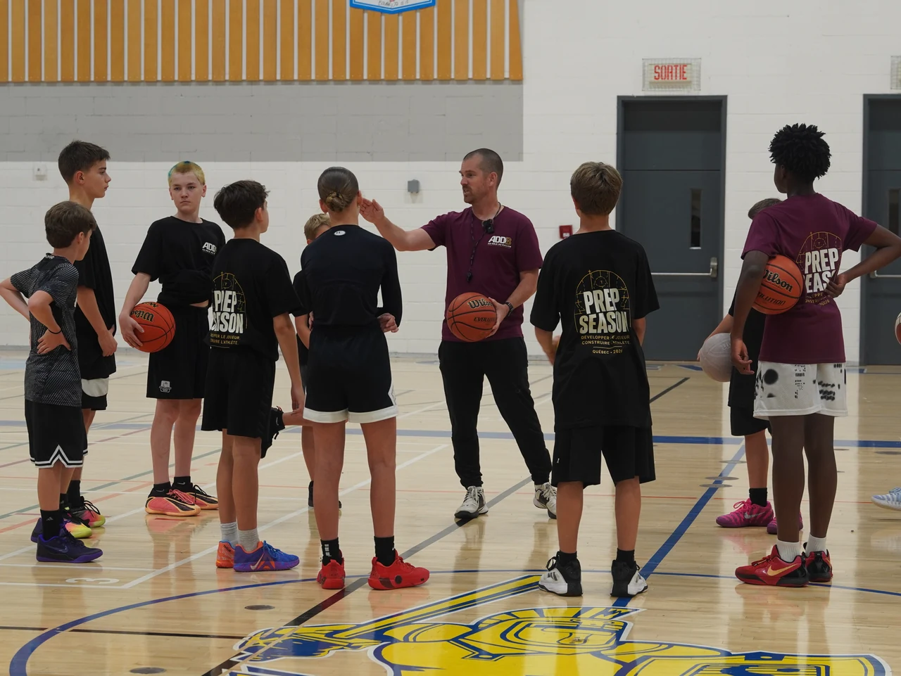 Charles Dubé-Brais, en chandail rouge ADDB, expliquant un exercice à un groupe de jeunes avec leurs ballons sur le terrain de basketball.